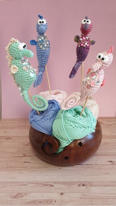Vier gehäkelte Amigurumi-Seepferdchen in Pastellfarben mit Perlen und Pailletten auf Holzstäbchen in einer Holzgarnschale.