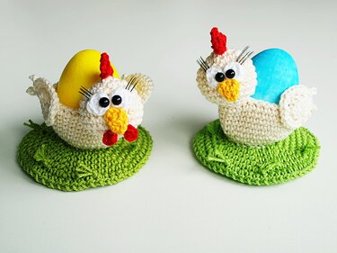 Zwei gehäkelte Hühner-Eierwärmer mit gelbem und blauem Ei auf grünen Häkelunterlagen