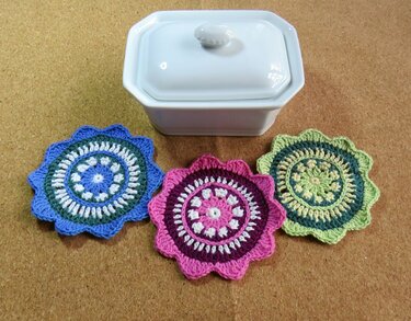 Drei gehäkelte Untersetzer in Blau, Pink und Grün auf einem Korkuntergrund neben einer weißen Schale