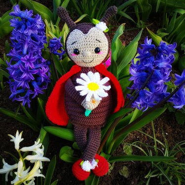 Lächelnde gehäkelte Marienkäfer-Puppe mit Gänseblümchen zwischen violetten Frühlingsblüten