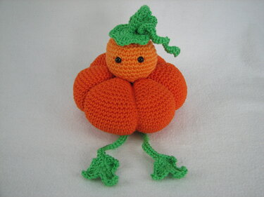Gehäkelter orangefarbener Kürbis-Amigurumi mit schwarzen Augen und grünen Blättern auf weißem Hintergrund
