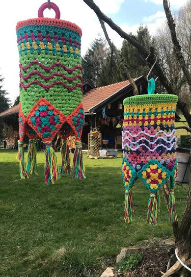 Zwei bunte, gehäkelte Lampions mit Fransen und Granny-Motiven hängen an einem Ast im Garten