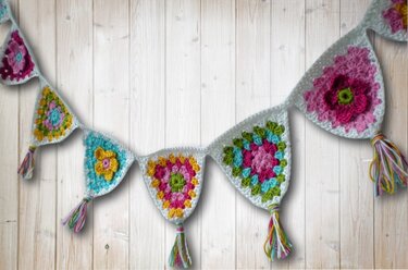 Gehäkelte Wimpelkette aus bunten Granny-Dreiecken mit weißem Rand und Quasten an einer Holzlattenwand