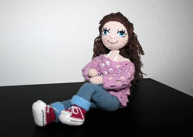Handmade amigurumi crochet doll with brown hair and blue eyes wearing a purple sweater, blue pants, and red-and-white sneakers, seated