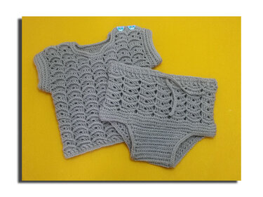 Grau gehäkeltes Baby-Set mit Muschelmuster, kurzärmeliges Oberteil mit zwei blauen Bärenknöpfen und passende kurze Hose auf gelbem Hintergrund.