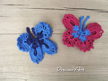 Zwei gehäkelte Schmetterlingsapplikationen, eine blau und eine pink, auf Holzuntergrund