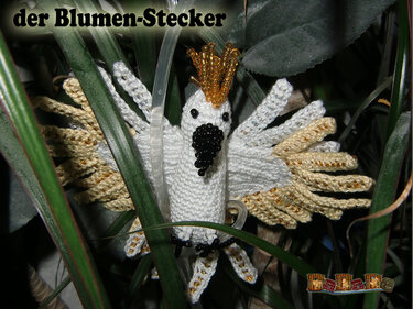 Gehäkelter weißer Kakadu-Blumenstecker mit goldfarbenen Perlen am Kamm und an den Flügeln zwischen grünen Pflanzenblättern.