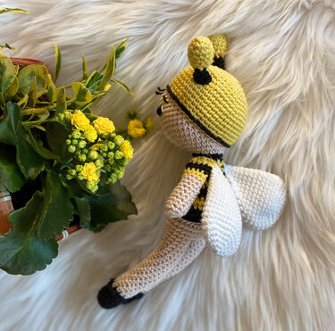 Gehäkelte Amigurumi-Biene in Gelb und Schwarz mit weißen Flügeln neben gelben Blüten auf hellem Fell