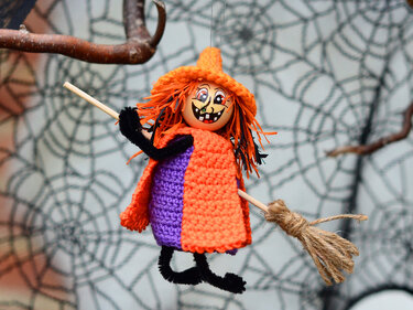 Gehäkelte Halloween-Hexe mit orangefarbenem Umhang und Besen vor Spinnennetz-Dekoration