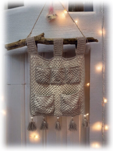 Gehäkelter Boho-Hängeorganizer mit Taschen und Quasten an einem Holzast, im Hintergrund Lichterketten.