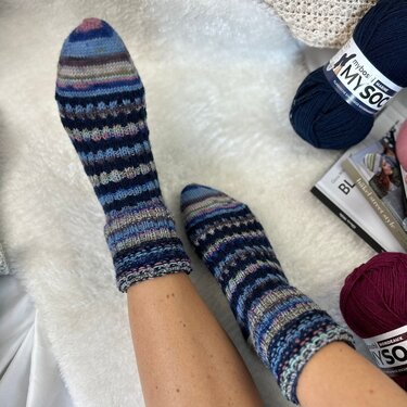 Füße in gestrickten, mehrfarbigen Socken auf weißem Flauschteppich, Wollknäuel im Hintergrund