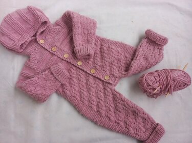 Altrosa gestrickter Baby-Overall mit Kapuze, Knöpfen und Zopfmuster, daneben Wollknäuel und Stricknadeln