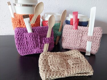 Mehrere gehäkelte Becherhüllen in Orange, Violett, Grün, Rosa und Beige, jede mit einer kleinen Tasche, die Holzlöffel und Zucker hält.
