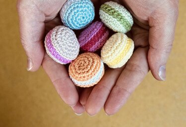 Offene Hände halten sechs kleine, gehäkelte Macarons in Pastellfarben