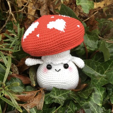 Gehäkelter Fliegenpilz mit rotem Hut und weißen Punkten, lächelnd, auf Efeu und Laub