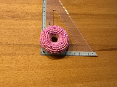 Kleiner rosa gehäkelter Donut neben transparentem Dreiecklineal auf Holz