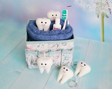 White knitted tooth amigurumi keychains with smiling faces displayed in a fabric box with a blue towel, toothbrush, and small bottle.