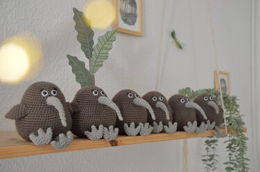 Sechs gehäkelte braune Amigurumi-Kiwis mit hellgrauen Schnäbeln und Füßen auf einem Holzregal.