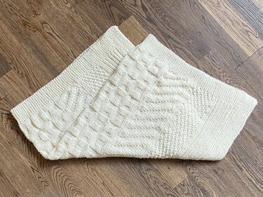 Gestrickte Babydecke in Creme mit verschiedenen Mustern, gefaltet auf Holzboden