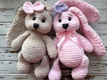 Two crocheted amigurumi bunnies in beige and pink with bows and a satin neck ribbon.
