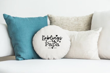 Rundes cremefarbenes Kissen mit schwarzem Schriftzug „Lieblings Papa“ und Herzmotiven auf einem Sofa.