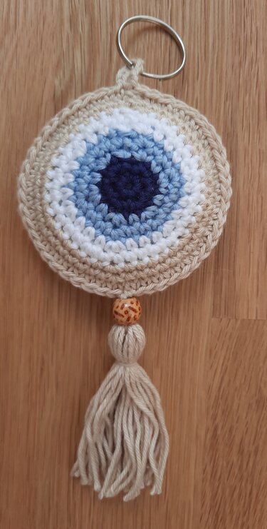 Runder gehäkelter Schlüsselanhänger mit Nazar‑Motiv in Blau, Weiß und Beige, Metallring, Holzperle und Quaste auf Holzuntergrund.
