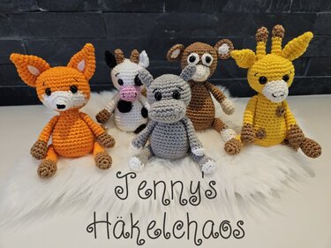 Fünf gehäkelte Amigurumi-Tiere (Fuchs, Kuh, Nilpferd, Affe, Giraffe) sitzend auf weißem Fell vor dunklem Hintergrund