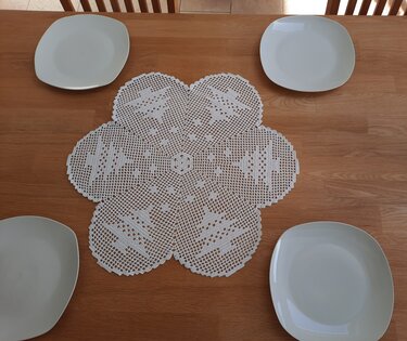 weißes blumenförmiges Häkeldeckchen mit sechs Lappen und Tannenbaum-Motiven, auf Holztisch gelegt, vier weiße Teller rundherum
