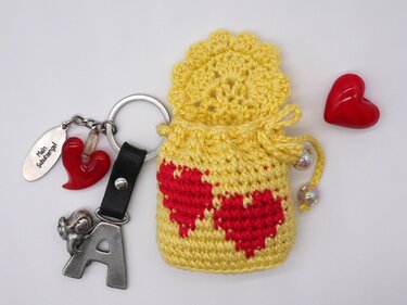 Gelbes handgehäkeltes Säckchen mit zwei roten Herz-Applikationen, Kordelzug und Schlüsselanhänger mit Buchstaben-Anhänger, Engel und ‚Mein Schutzengel‘-Anhänger auf weißem Hintergrund.