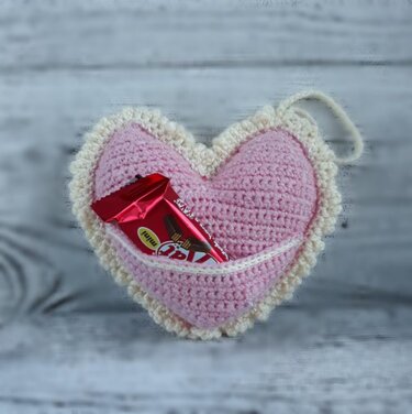 Pink crocheted heart with cream ruffled edge and front pocket holding a small wrapped treat.