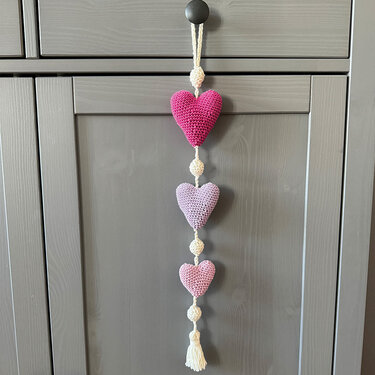 Drei hängende, gehäkelte Pastellherzen an einer Kordel mit Perlen und Quaste, hängend an einem Schrankknauf.