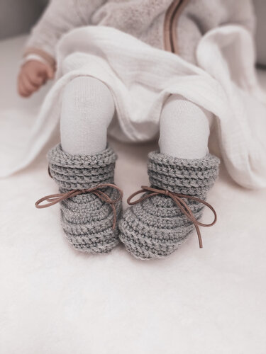 Gehäkelte graue Babyschuhe mit braunen Schnürbändern an Babyfüßen auf hellem Stoff
