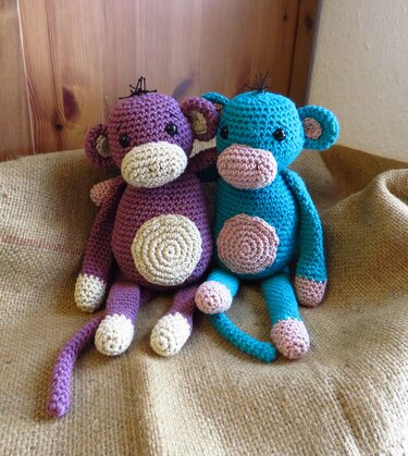 Zwei gehäkelte Amigurumi-Affen, einer lila und einer türkis, sitzen auf Jutestoff vor Holzvertäfelung.