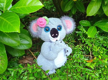 Gray crocheted koala amigurumi with big blue eyes and a pink flower on its head, sitting surrounded by green leaves