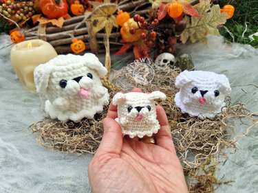 Hand holding a small white crocheted ghost dog amigurumi, with two matching amigurumi and Halloween decorations in the background.