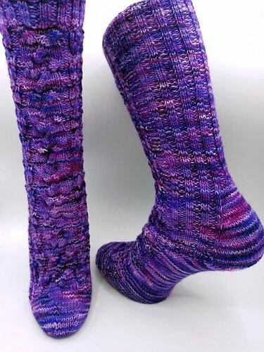 Zwei lila-melierte handgestrickte Socken mit strukturiertem Muster vor hellem Hintergrund, eine aufrecht, eine liegend.