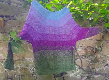 Weiter gestrickter Pullover mit Farbverlauf in Blau, Violett und Grün an einer Leine vor Steinmauer