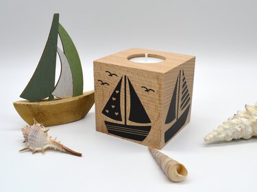 Quadratischer Holzteelichthalter mit schwarzem Segelbootmotiv, daneben eine kleine grüne Holzsegelbootfigur und Muscheln.