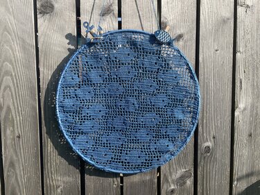 Rundes blaues Filethäkel-Wandbild in rundem Rahmen an einer Holzwand mit kleinen Holzanhängern