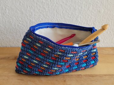 Gehäkelte dunkelblaue Reißverschlusstasche mit bunten Akzenten und offenem Innenraum, darin Häkelnadeln auf einem Holztisch