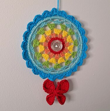 Gehäkelter runder Traumfänger in Regenbogenfarben mit facettiertem Kristall in der Mitte und rotem Schmetterling an weißer Wand