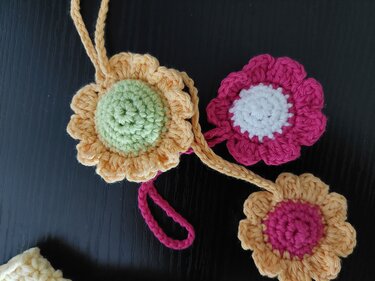 Drei gehäkelte Blüten in unterschiedlichen Farbkombinationen an einer gehäkelten Kordel auf schwarzem Hintergrund.