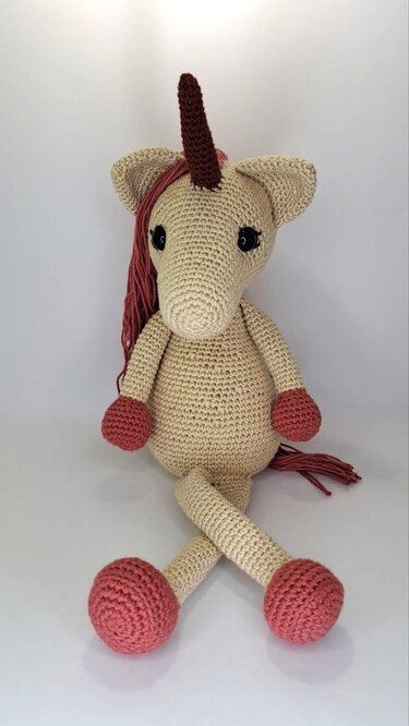 Gehäkeltes Amigurumi-Einhorn in Beige mit dunkelbraunem Horn, bordeauxroter Mähne und Hufen, sitzend mit überkreuzten Beinen