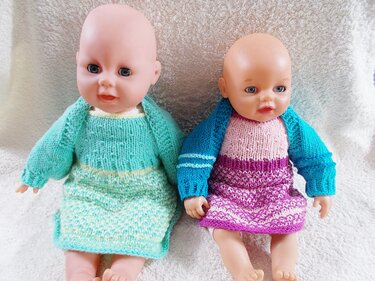 Zwei Babypuppen in gestrickten Kleidern und Boleros, links mint, rechts rosa-lila