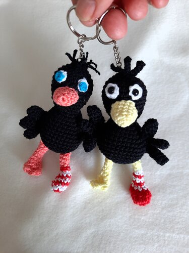 Zwei gehäkelte schwarze Vogel‑Amigurumi als Schlüsselanhänger mit bunten Schnäbeln und gestreiften Socken an Metallringen