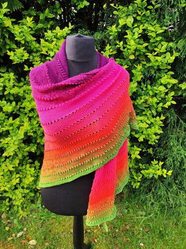 Gestricktes Dreieckstuch mit Farbverlauf in Pink, Orange und Grün auf schwarzer Puppe vor grüner Hecke