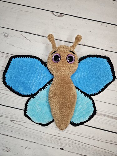 Gehäkelter Amigurumi‑Schmetterling mit hellblauen und dunkelblauen Flügeln, braunem Körper und schwarzen Randkanten auf hellen Holzlatten