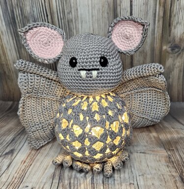 Gehäkelte graue Fledermaus-Amigurumi mit rosa Ohren und leuchtendem, durchbrochenem Bauch auf Holzuntergrund.