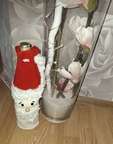 Gehäkelte Santa‑Flaschenhülle mit roter Mütze, weißem Bart und Knopf‑Nase neben einer Glasvase