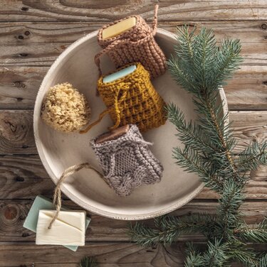 Drei gestrickte Seifentaschen in Holzschale neben Seife, Schwamm und Tannenzweig